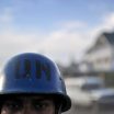 Close up on a brown-skinned man wearing a blue helmet with "UN" stamped on it in black letters. Only the man's eyes and the top of his head are visible. The background is blurred.