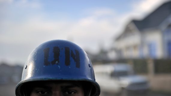Close up on a brown-skinned man wearing a blue helmet with "UN" stamped on it in black letters. Only the man's eyes and the top of his head are visible. The background is blurred.