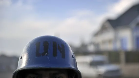 Close up on a brown-skinned man wearing a blue helmet with "UN" stamped on it in black letters. Only the man's eyes and the top of his head are visible. The background is blurred.