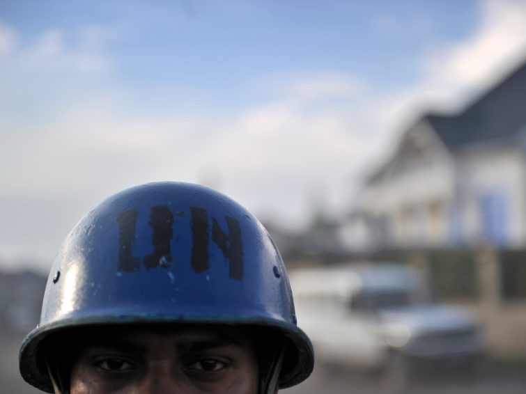 Close up on a brown-skinned man wearing a blue helmet with "UN" stamped on it in black letters. Only the man's eyes and the top of his head are visible. The background is blurred.